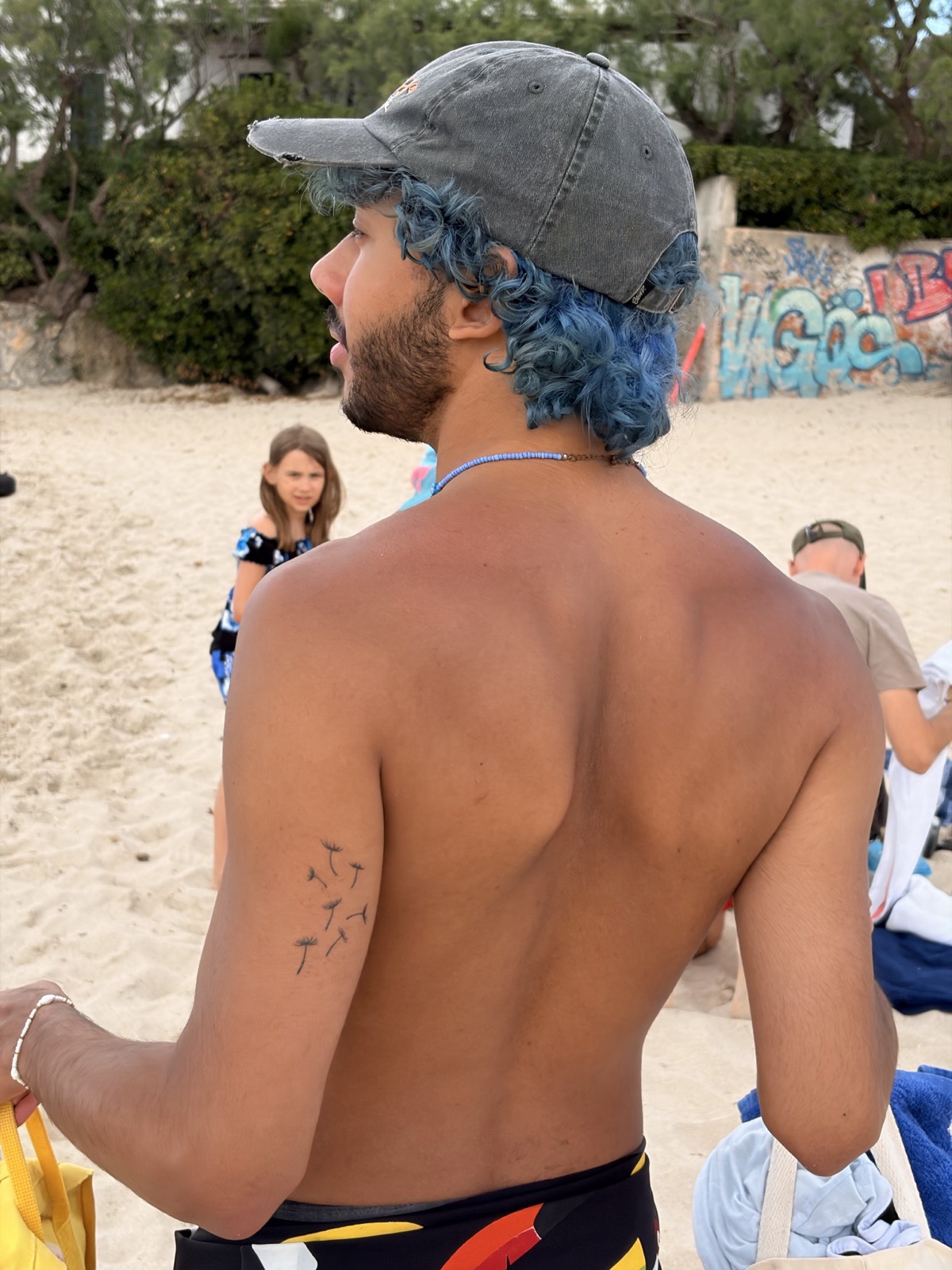 Mat photographed from behind on a beach, with blue curly hair under a cap
