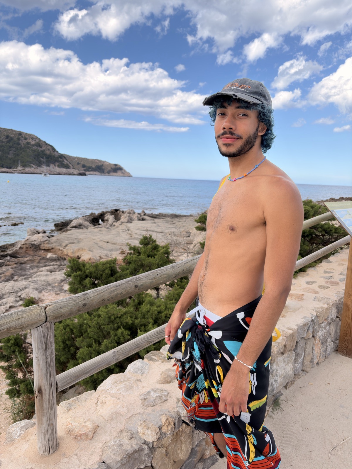 Mat on a stone path by the sea, wearing a cap and a colourful sarong